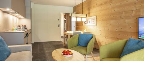 A modern living room with a dining area, wooden wall paneling, a television, and a bowl of fruit on a small table.