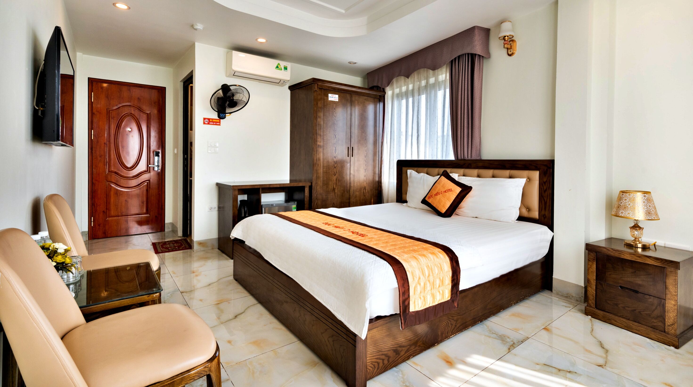 A hotel room with a large bed, a wooden wardrobe, a desk, a chair, a TV, and a lamp.