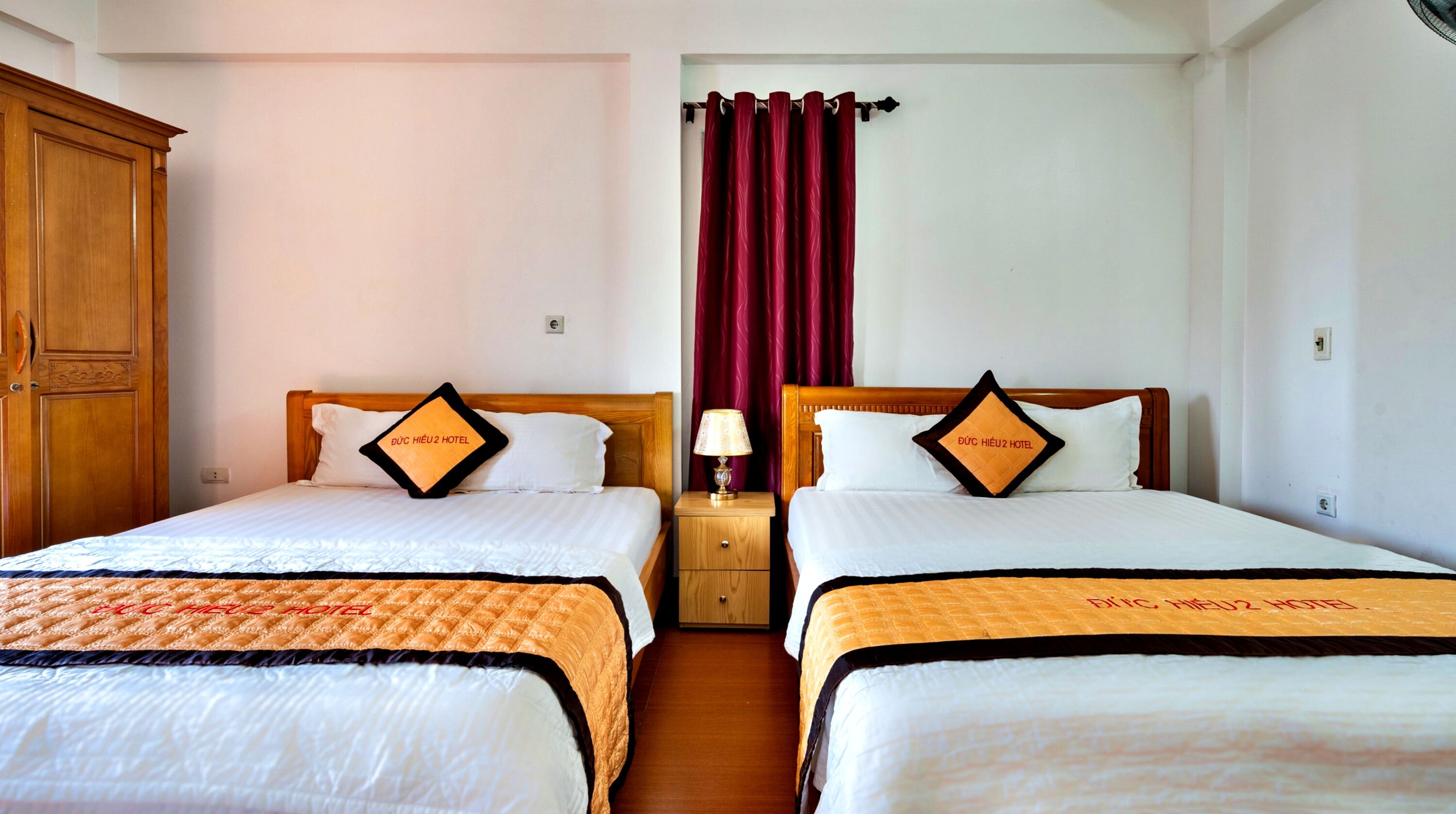 A hotel room with two beds, each with a matching pillow and bedspread, a wooden wardrobe, a nightstand with a lamp, and a red curtain.
