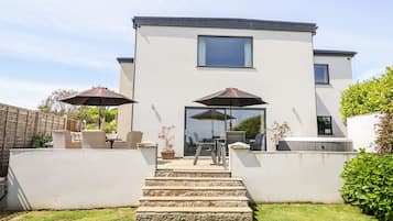 Ein modernes, zweistöckiges Haus mit einer Terrasse, die Gartenmöbel und Sonnenschirme bietet.