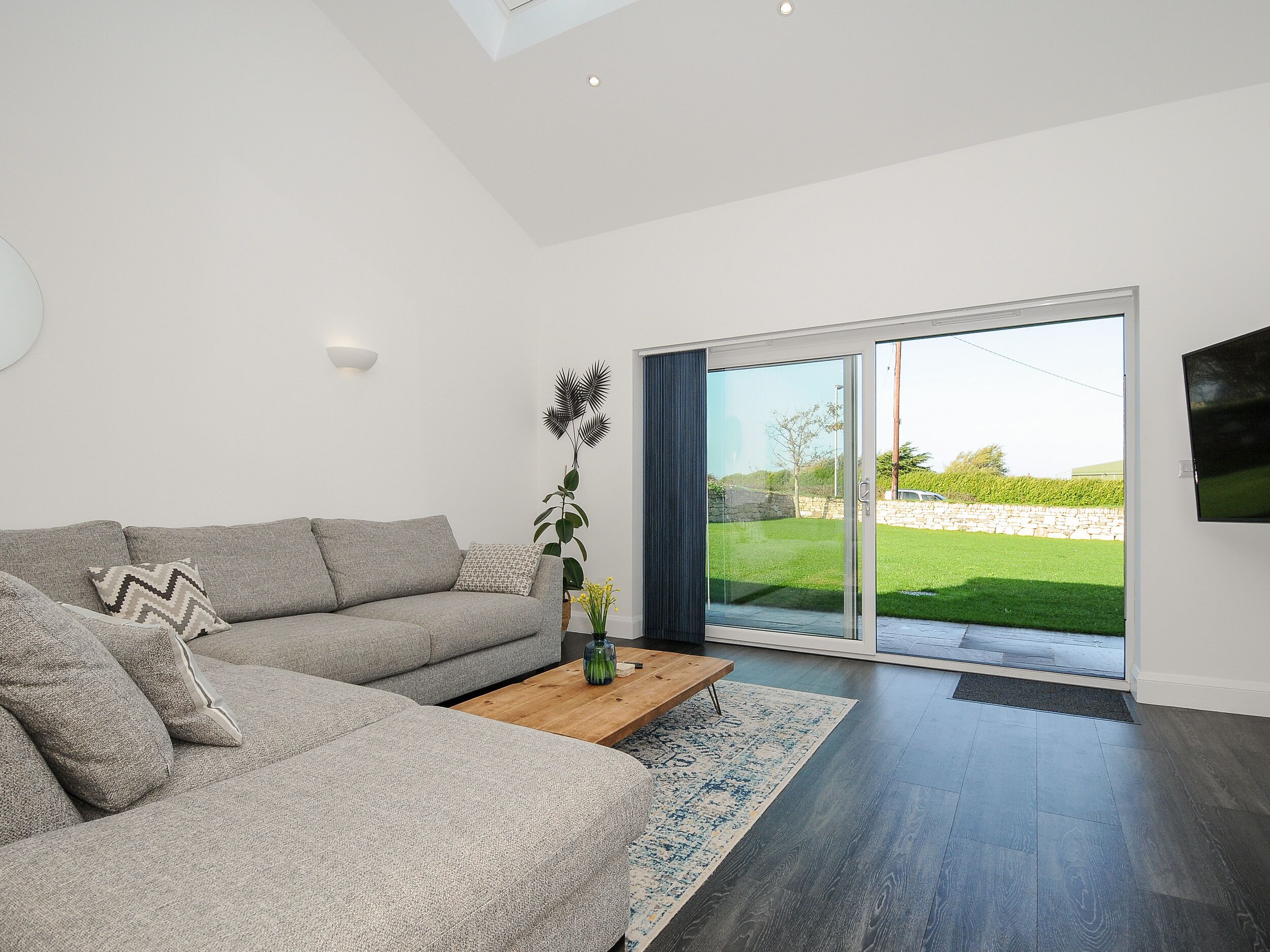 A modern living room with a grey sectional sofa, a wooden coffee table, and a large sliding glass door leading to a garden.