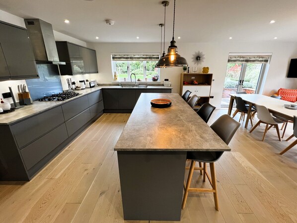 A modern kitchen with a central island, dark cabinetry, and a large window providing a view of greenery. - Laurel Cottage (Newchurch)
