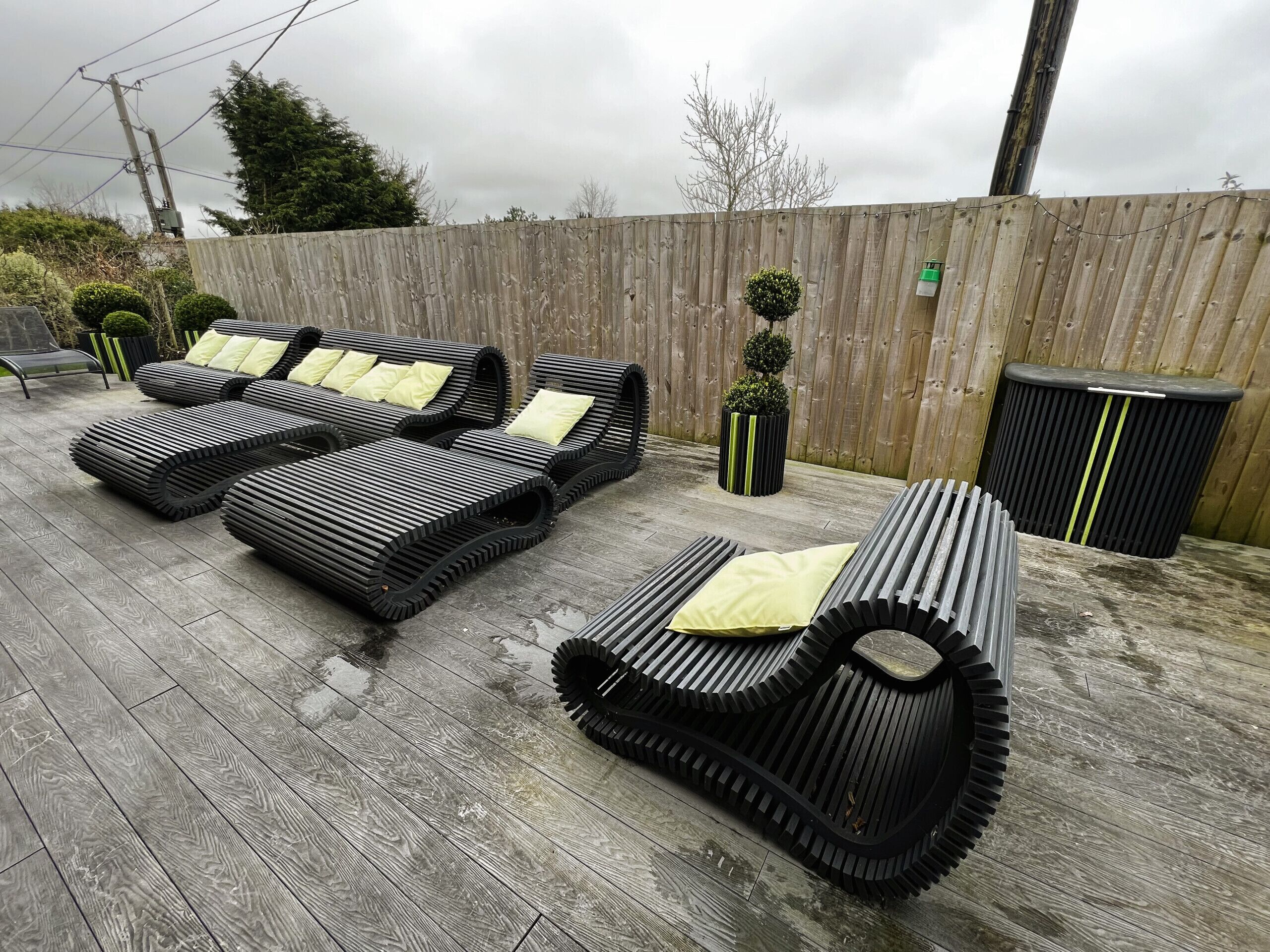A patio area with three black lounge chairs with yellow cushions, a small potted plant, and a trash bin.