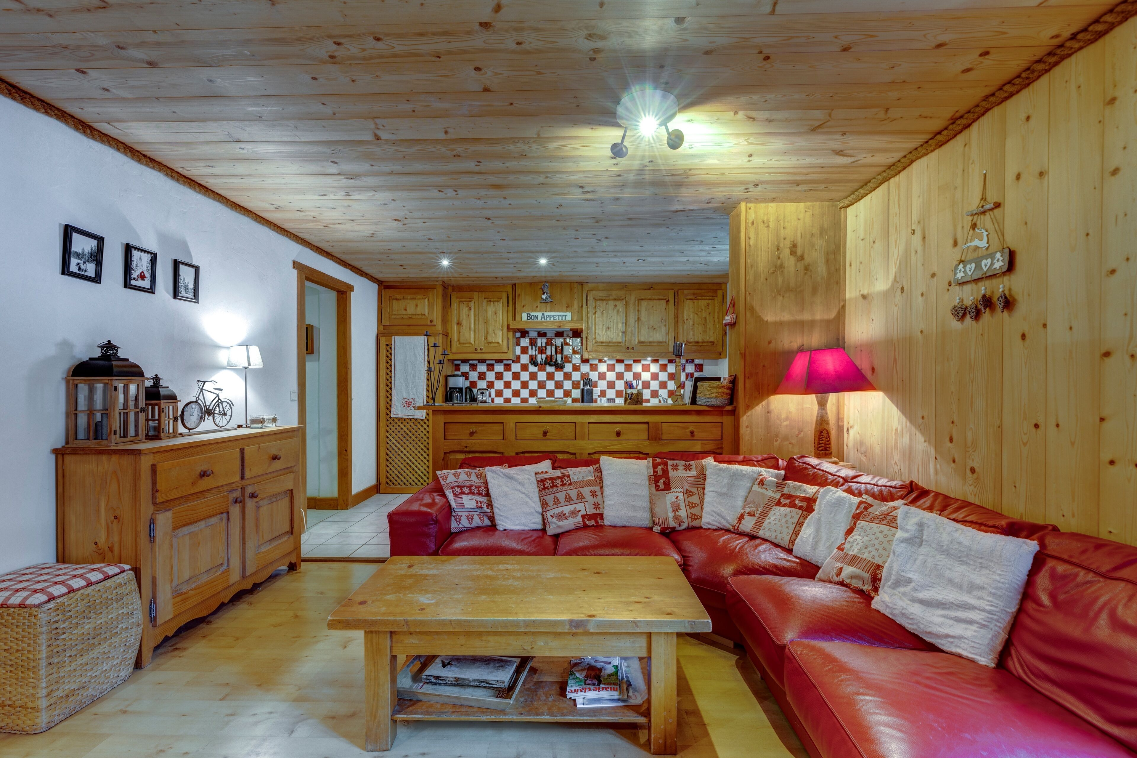 Un salon chaleureux avec un canapé en cuir rouge, une table basse en bois et un coin cuisine doté de meubles en bois et d’un pare-feu à carreaux.