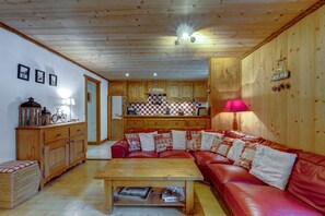 Un salon chaleureux avec un canapé en cuir rouge, une table basse en bois et un coin cuisine doté de meubles en bois et d’un pare-feu à carreaux.