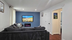 A living room with a large flat-screen TV mounted on a blue wall, a gray sofa, and a fireplace. - Short Walk to Metro - Fenced Yard + Covered Patio (Silver Spring)