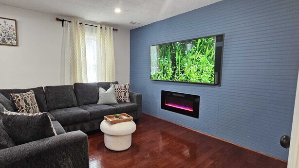 A modern living room with a sectional sofa, a white pouf, a flat-screen TV mounted on a blue wall, and a fireplace below it. - Short Walk to Metro - Fenced Yard + Covered Patio (Silver Spring)