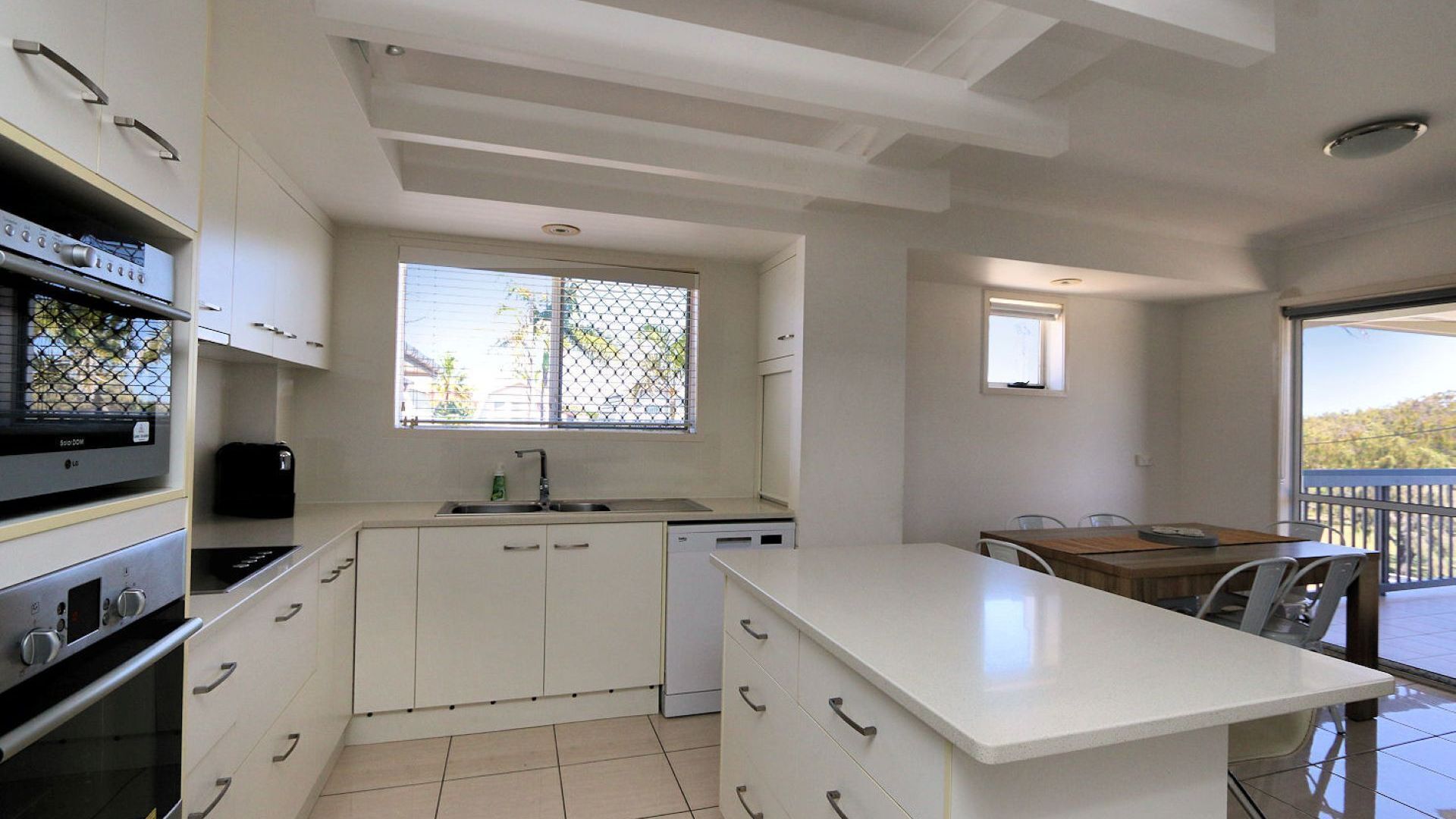 Une cuisine moderne avec des meubles blancs, un four encastré et un lave-vaisselle. On y trouve un coin repas avec une table et des chaises, ainsi qu’un balcon doté d’une rambarde.