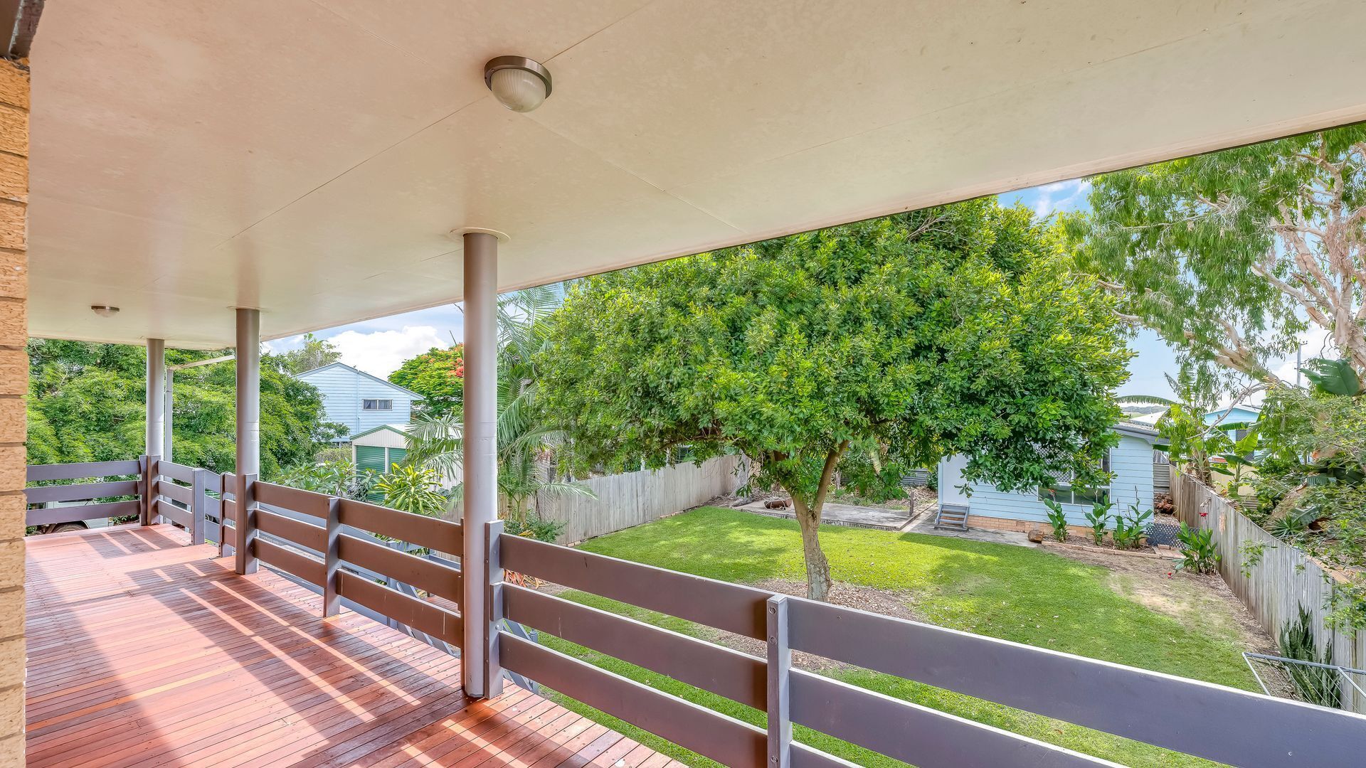 A covered patio area with a wooden deck and metal railings, overlooking a backyard with a lawn and trees.