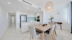 A modern kitchen with white cabinetry, a central island, and a dining area with a wooden table and chairs. - C Bargara Unit 101 Ocean Front (Bargara)