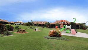 A well-maintained grassy area with picnic tables, a playground, and a building with a red-tiled roof.
