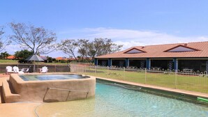 A swimming pool with a sunshade, a fenced area with lounge chairs, and a building with a tiled roof.