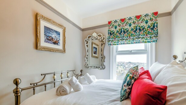 A neatly made bed with white linens and rolled towels, a large window with a view, and a decorative mirror on the wall.