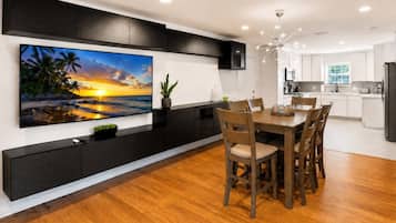 Una cocina moderna con armarios blancos, un comedor con mesa y sillas de madera, y un televisor de pantalla plana grande montado en la pared que muestra una escena de atardecer.