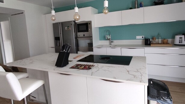 A modern kitchen with white cabinetry, a marble countertop, and a central island. There are hanging light fixtures, a microwave, and a refrigerator. - Cottage Chaleureux Proche de L'océan (Grayan-et-l'Hôpital)