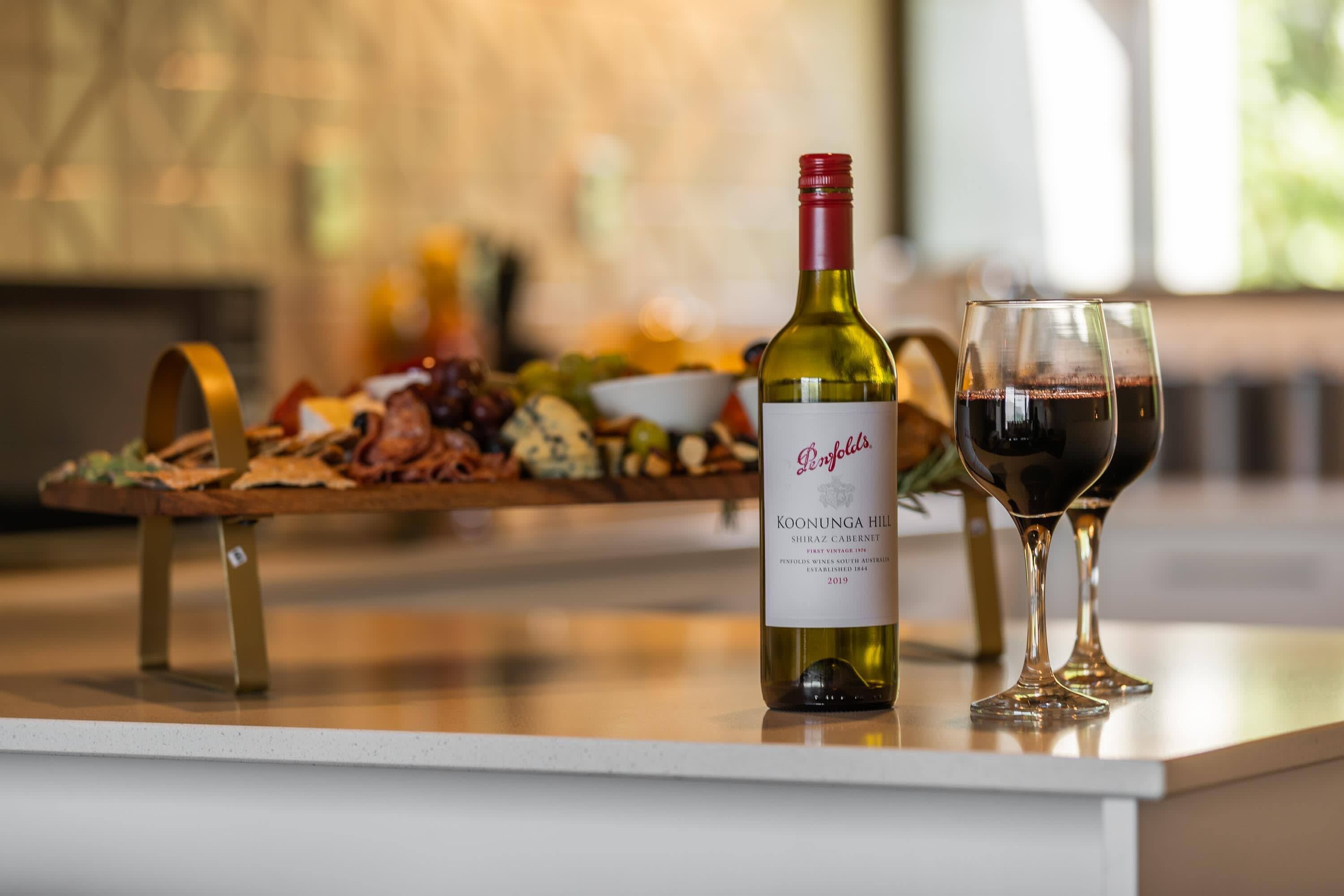 Pullo Koonunga Hill Shiraz Cabernet -punaviiniä, viinilasi sekä puinen tarjoilulauta, jossa on erilaisia juustoja ja leikkeleitä.