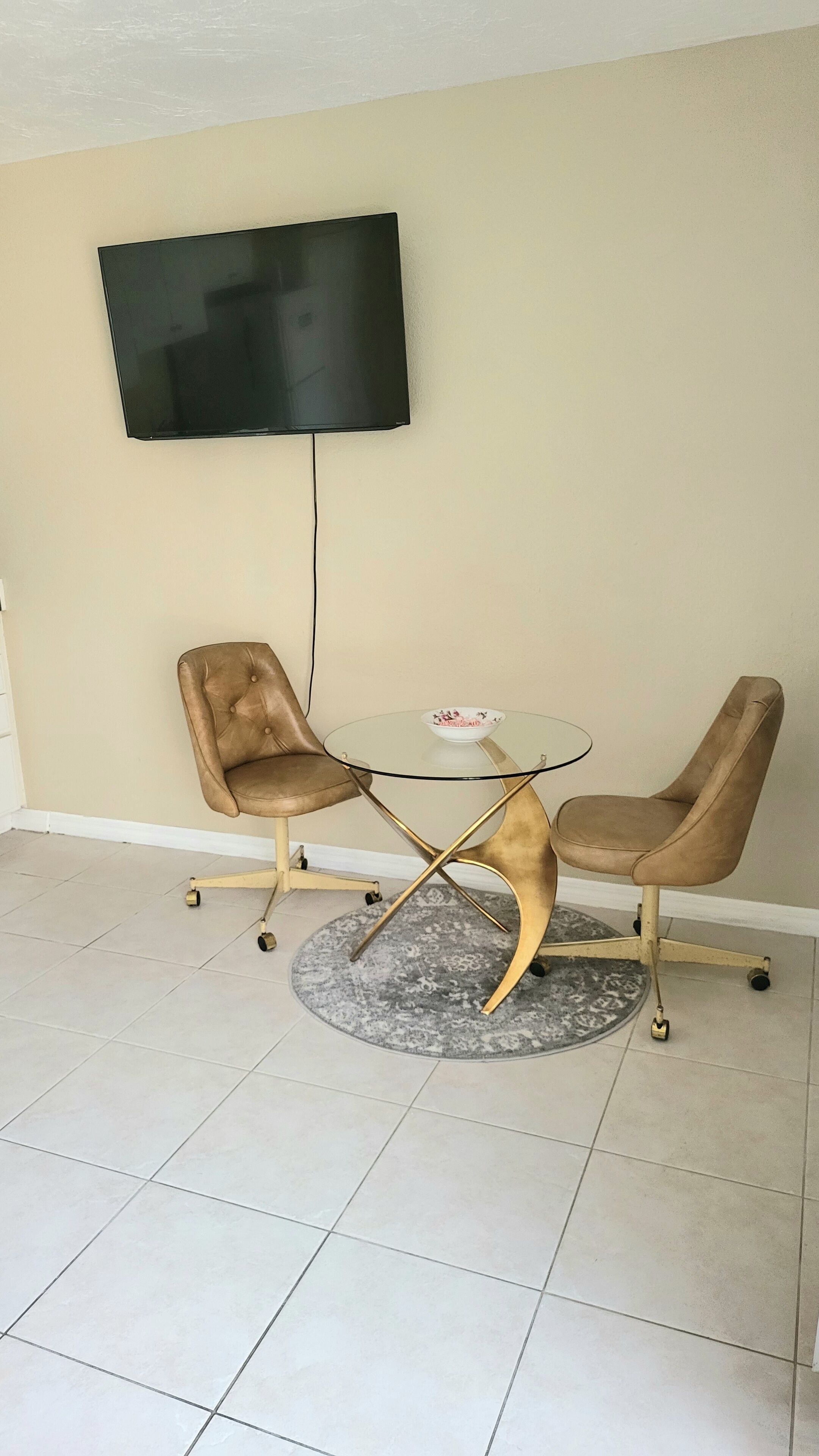 A small room with a TV mounted on the wall, two chairs, a glass table with a bowl, and a patterned rug.