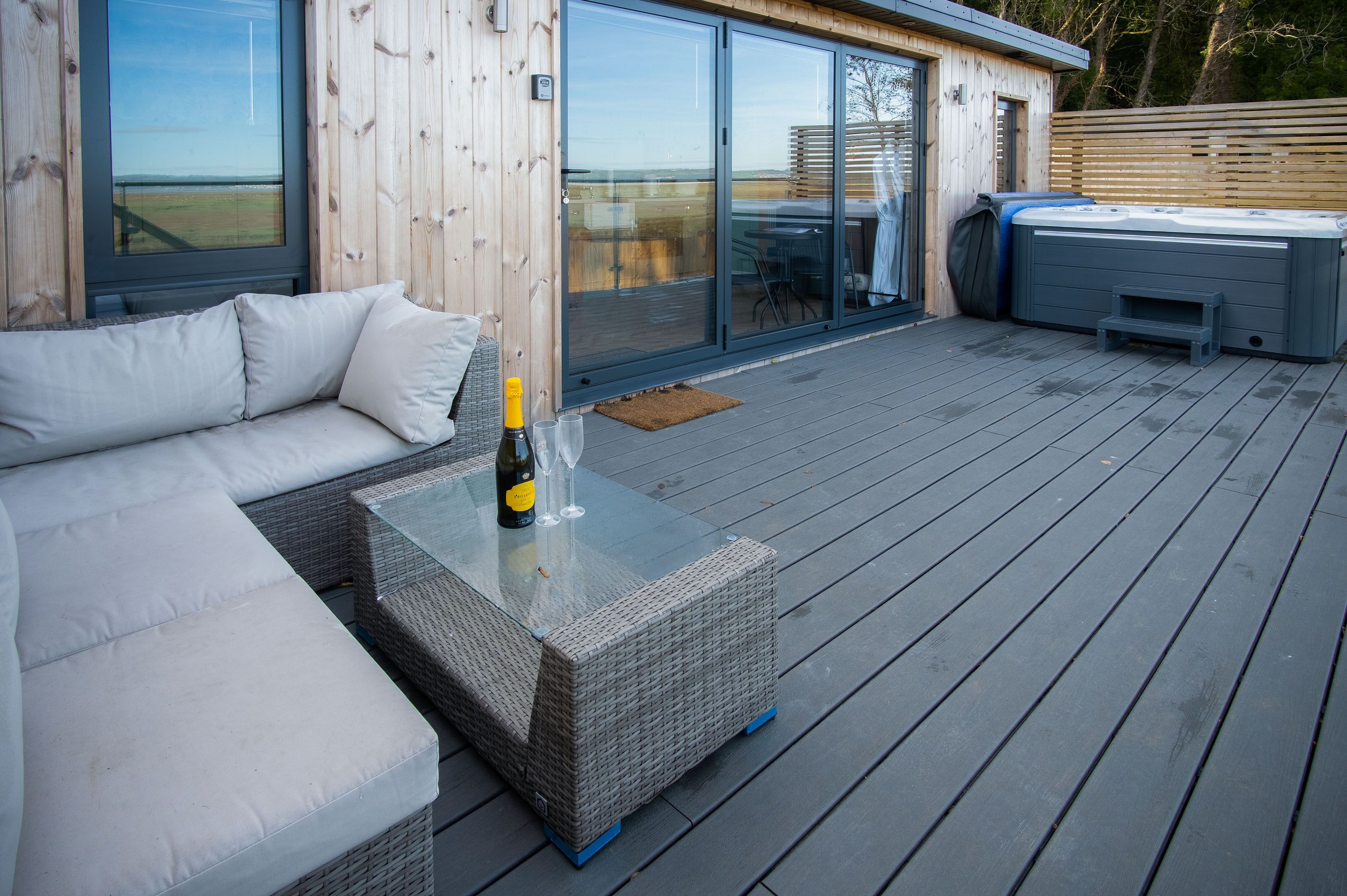 Une terrasse en bois avec une table à plateau de verre, une bouteille de champagne et un canapé blanc.