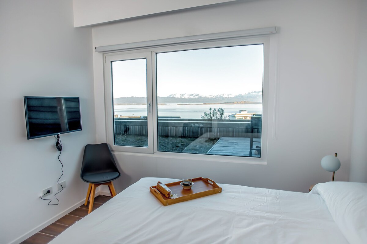 Habitación de hotel con cama, televisor montado en la pared, una silla, ventana con vistas a montañas y un lago, y una bandeja con una taza y un pequeño recipiente sobre la cama.