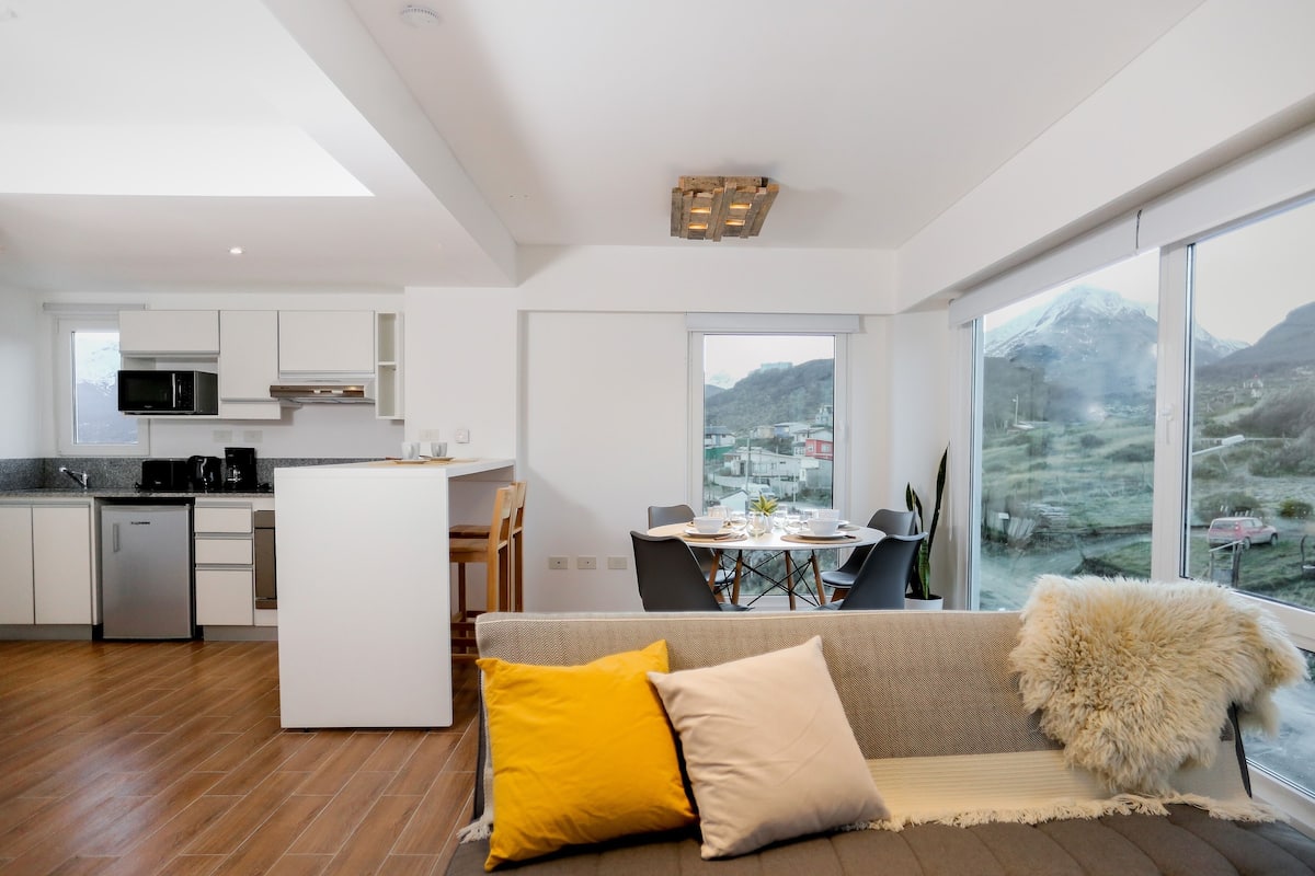 Cocina moderna con armarios blancos, microondas y lavavajillas.  Comedor con mesa y sillas, y sofá con cojines. Amplias ventanas ofrecen vista a un paisaje montañoso.