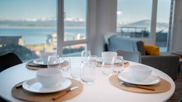 Mesa de comedor con platos, tazones y vasos blancos, con vista al mar y montañas al fondo.