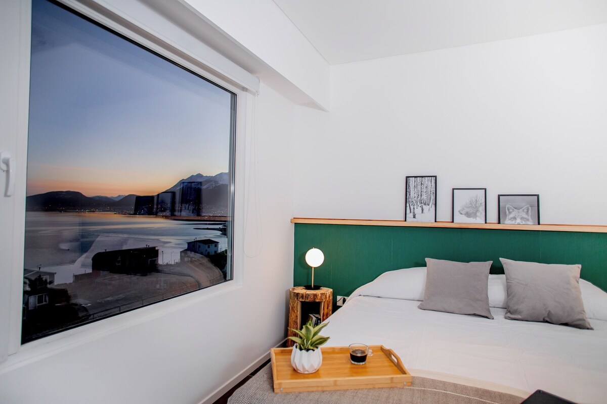 Quarto de hotel com uma janela grande, cama com cabeceira verde, mesa de cabeceira com abajur e quadros nas paredes.