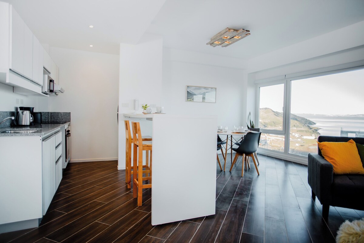 Uma cozinha moderna com piso de madeira escura, armários brancos e uma área de jantar com vista para o oceano.