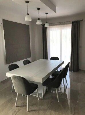 Un comedor moderno con una mesa larga, seis sillas, lámparas colgantes y un ventanal con cortinas.