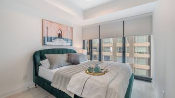 A modern bedroom with a green upholstered bed, a large window with blinds, a framed artwork above the bed, and a bedside table with a lamp.