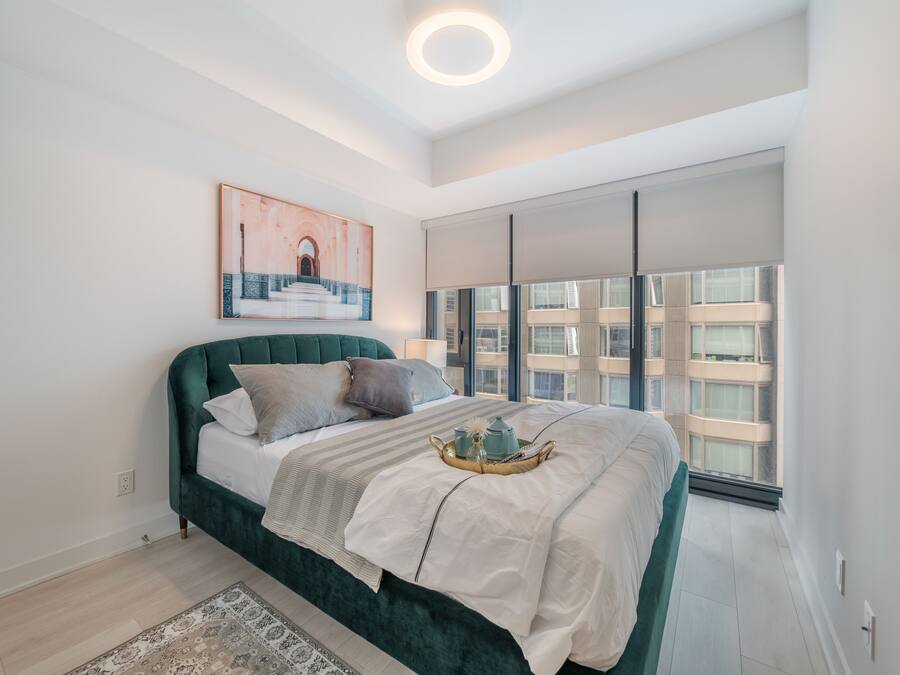 A modern bedroom with a green upholstered bed, a large window with blinds, a framed artwork above the bed, and a bedside table with a lamp.