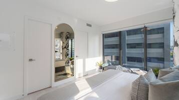 Un dormitorio moderno con una cama grande, un vestidor y vistas a un paisaje urbano a través de ventanales de gran altura.