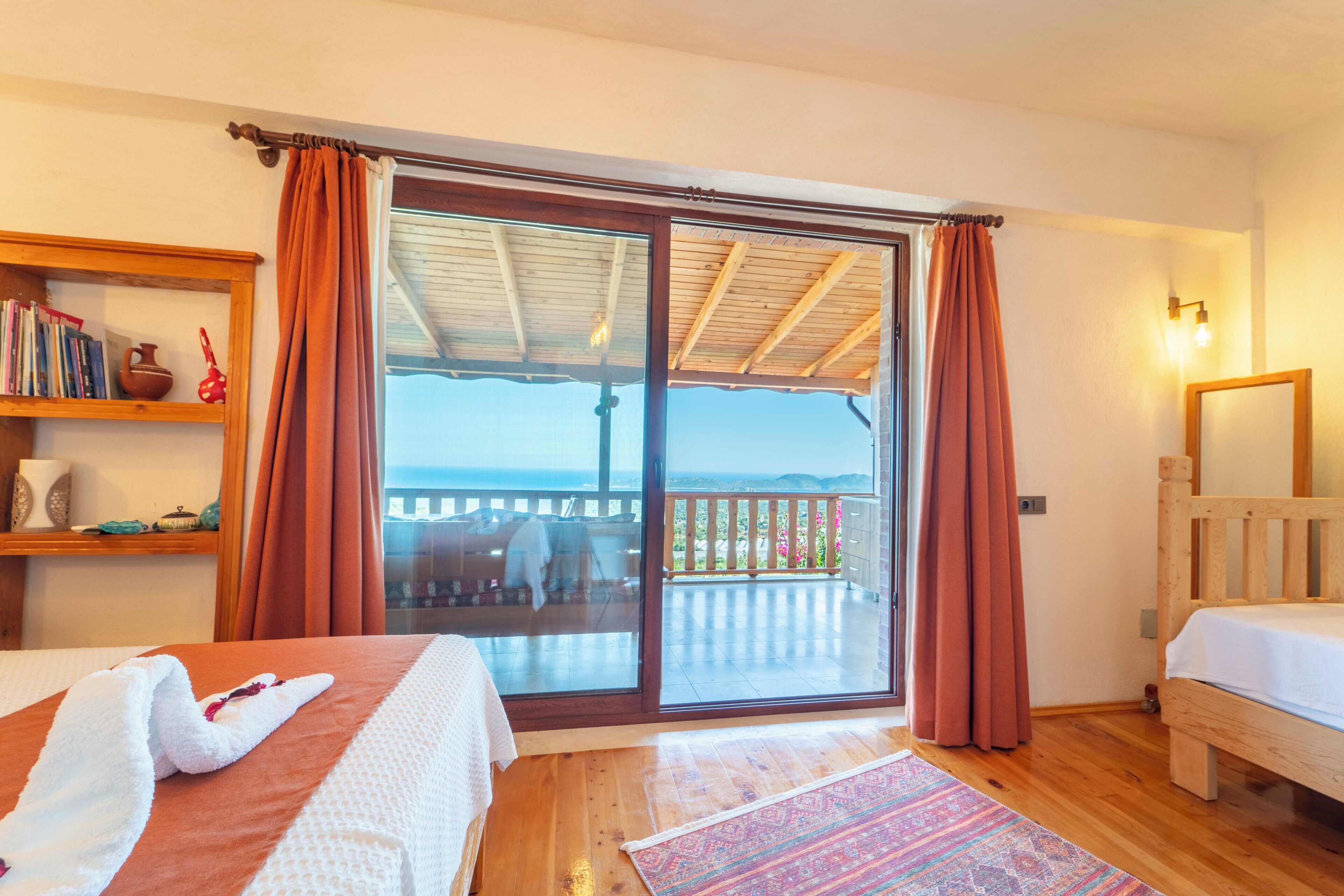 A bedroom with a large sliding glass door leading to a balcony, a wooden bed, a bedside table with books, and a nightstand.