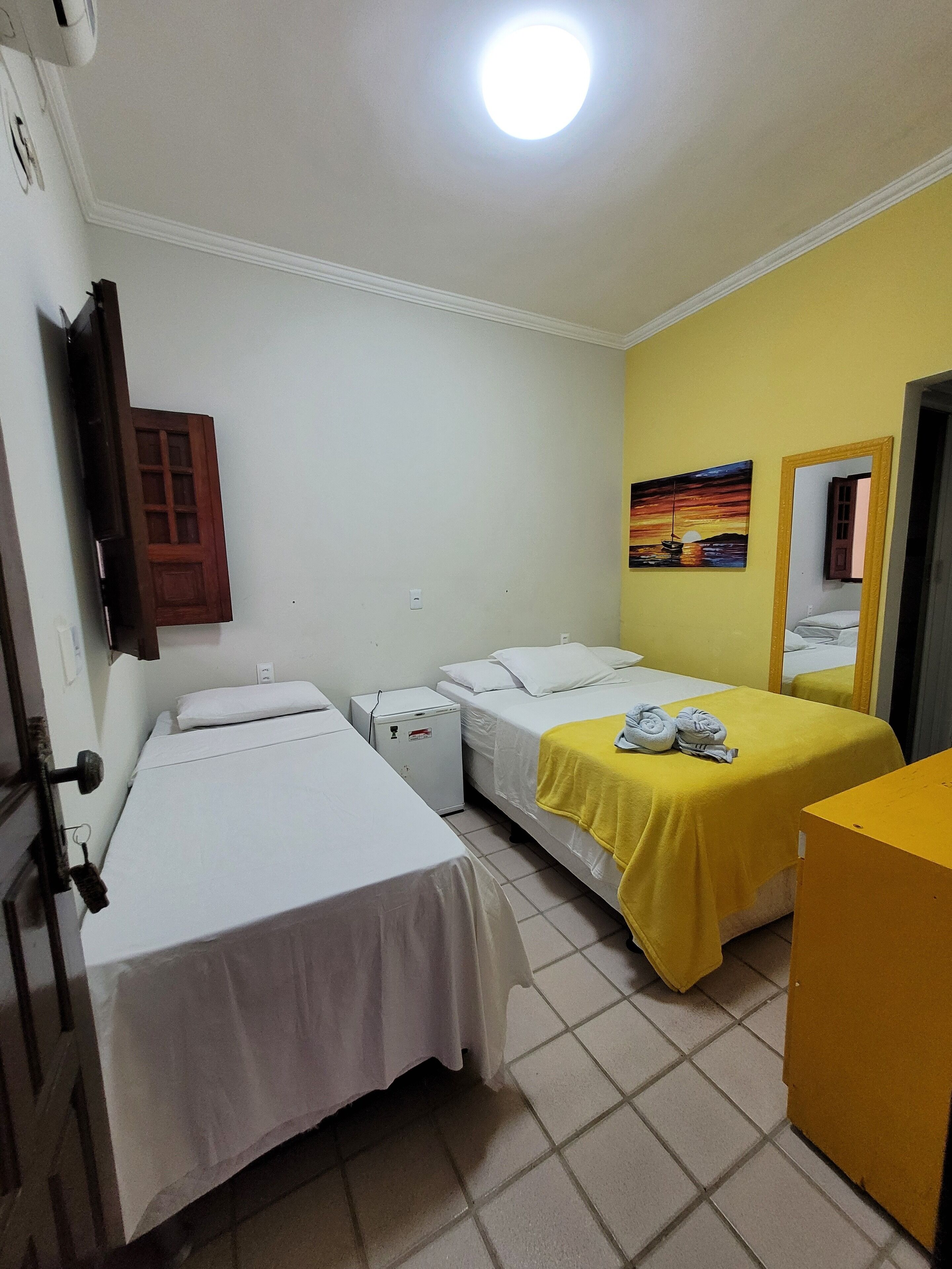 Ein Hotelzimmer mit zwei Einzelbetten, einem kleinen Kühlschrank, einem Nachttisch mit gellem Tuch sowie einem Bild an der Wand und einem Spiegel.