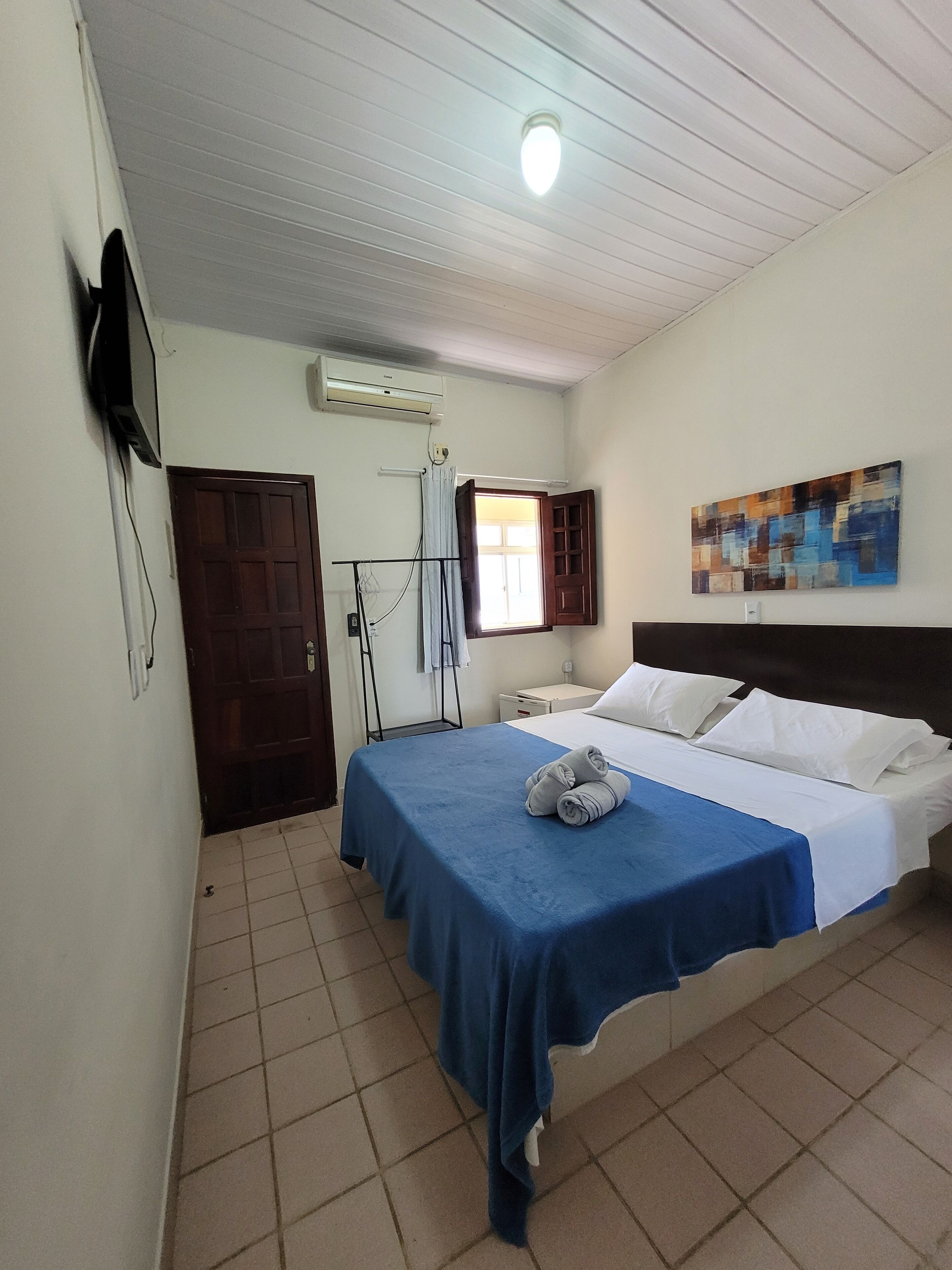 A hotel room with a bed, a flat-screen TV mounted on the wall, an air conditioning unit, a window with a curtain, and a painting on the wall.