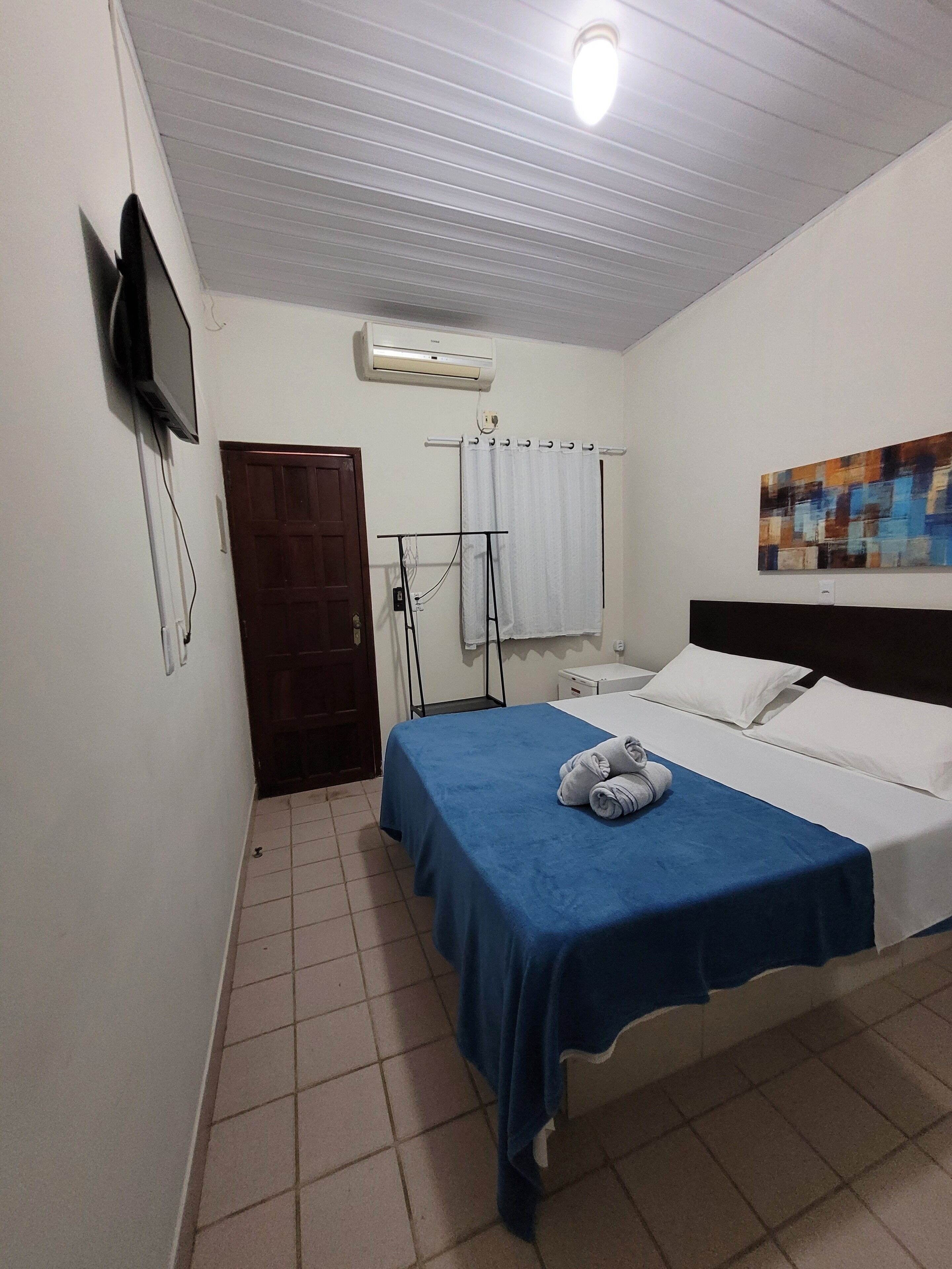 A hotel room with a bed, a TV mounted on the wall, an air conditioning unit, a curtain rod, and a painting on the wall.