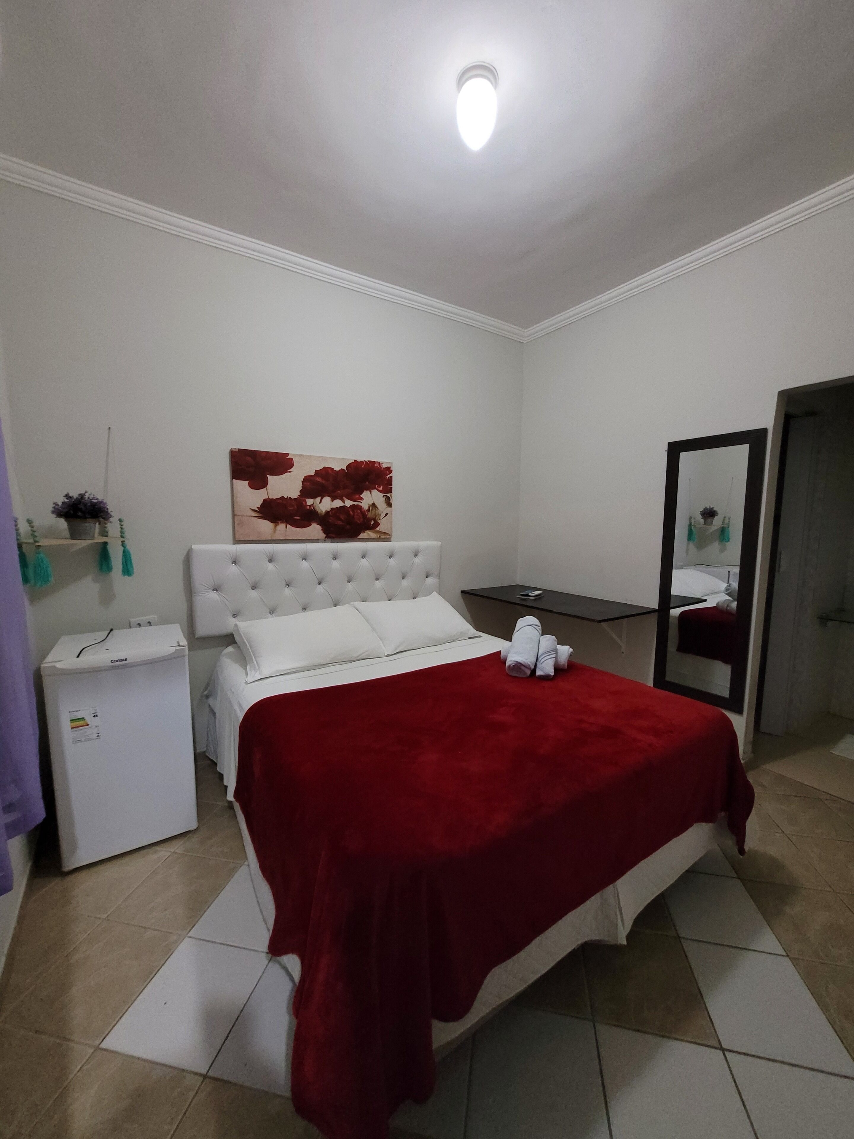 A bedroom with a white bed, red blanket, and a painting on the wall. There is a small white refrigerator and a mirror on the wall.