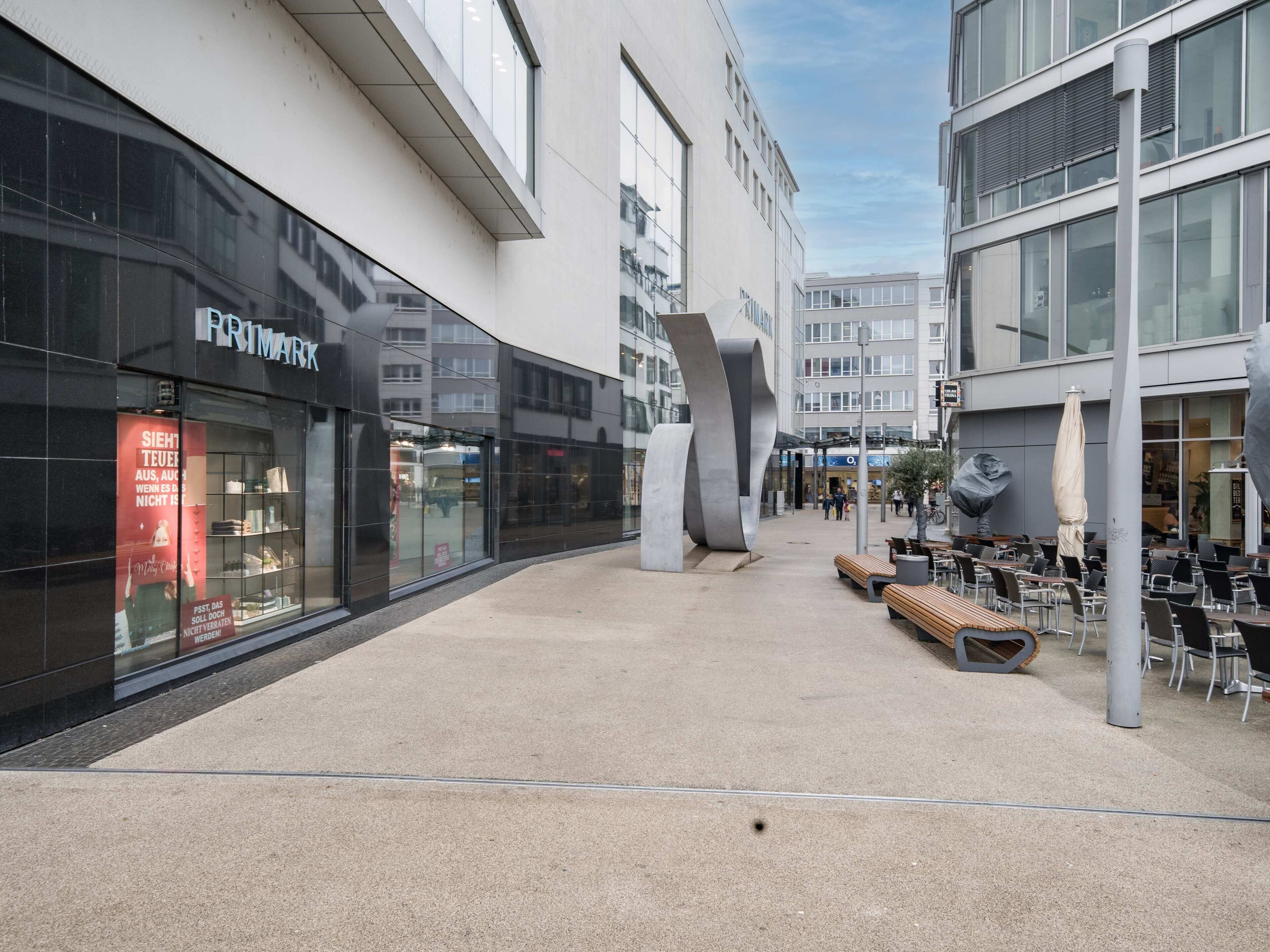 A modern urban plaza with a Primark store, outdoor seating, and a large abstract sculpture.