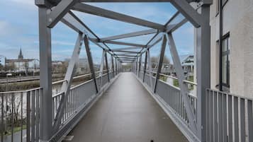 Eine Fußgängerbrücke mit Metallfachwerkkonstruktion, einer ebenen Gehweg und Blick auf Gebäude sowie eine Kirche im Hintergrund.