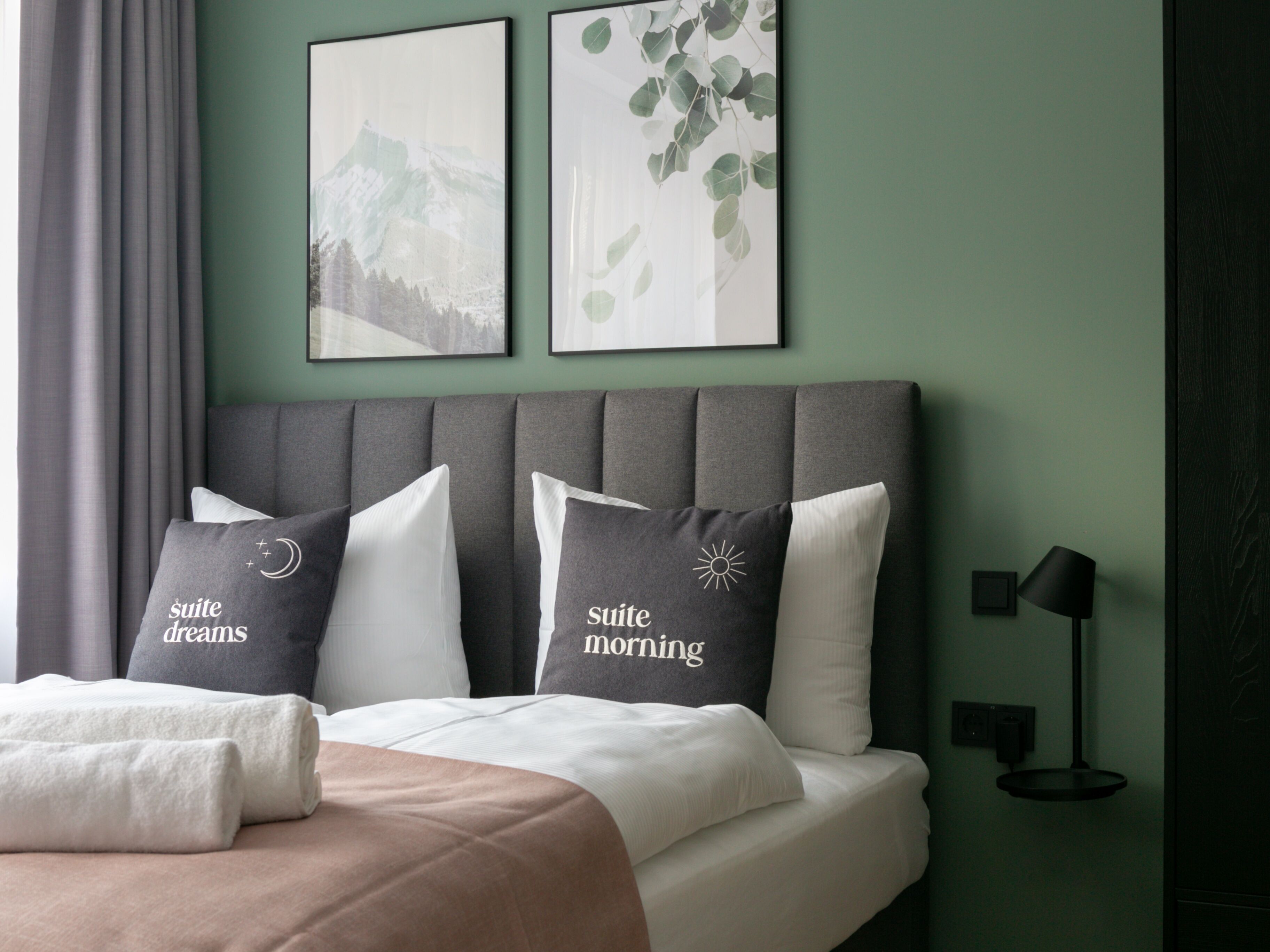 Una cama cuidadosamente arreglada con almohadas que dicen "suite dreams" y "suite morning" en una habitación con paredes verdes y un cabecero gris.
