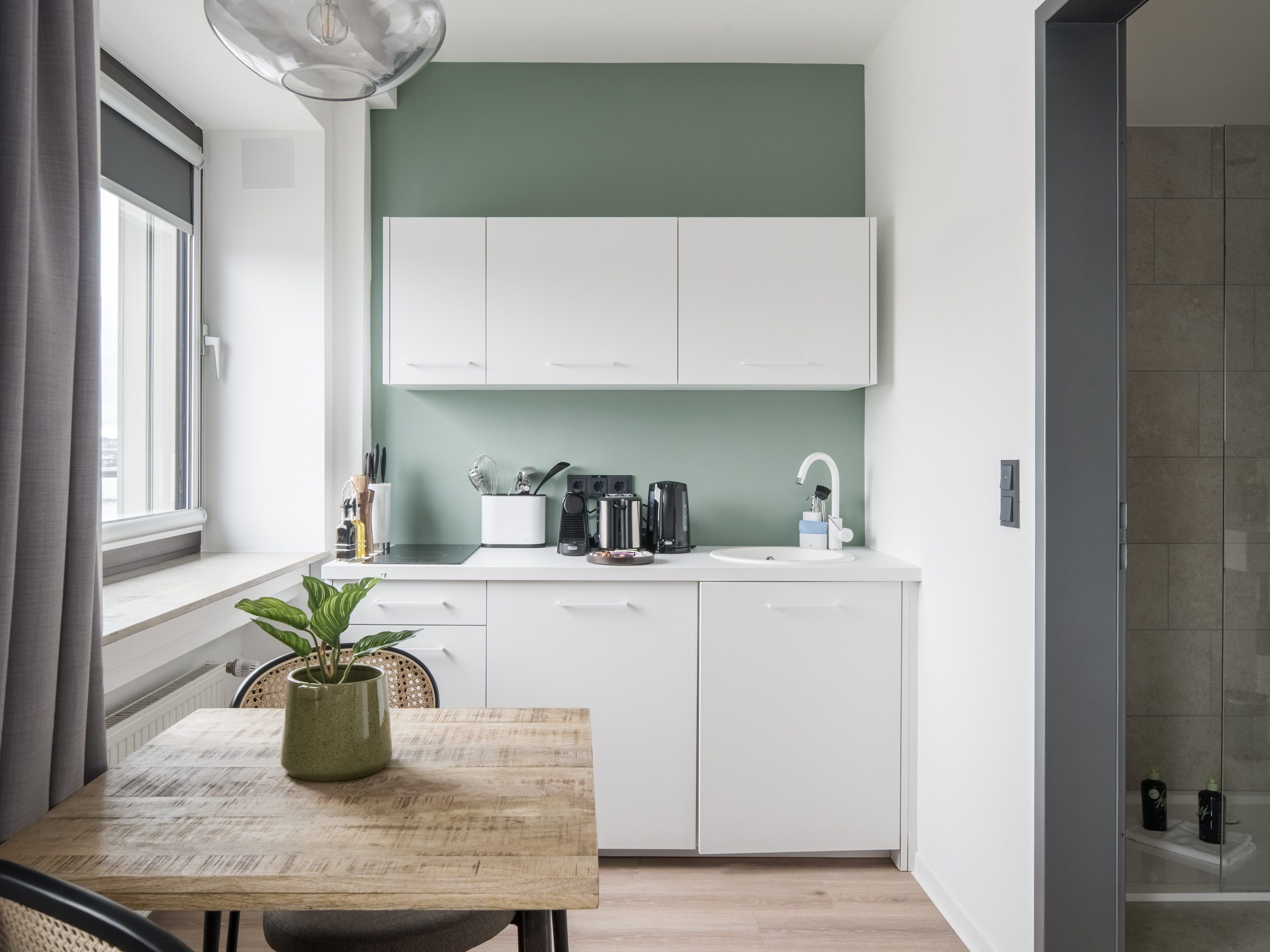 A compact kitchen with white cabinets, a coffee machine, and a sink. There's a dining table with a plant and a chair.