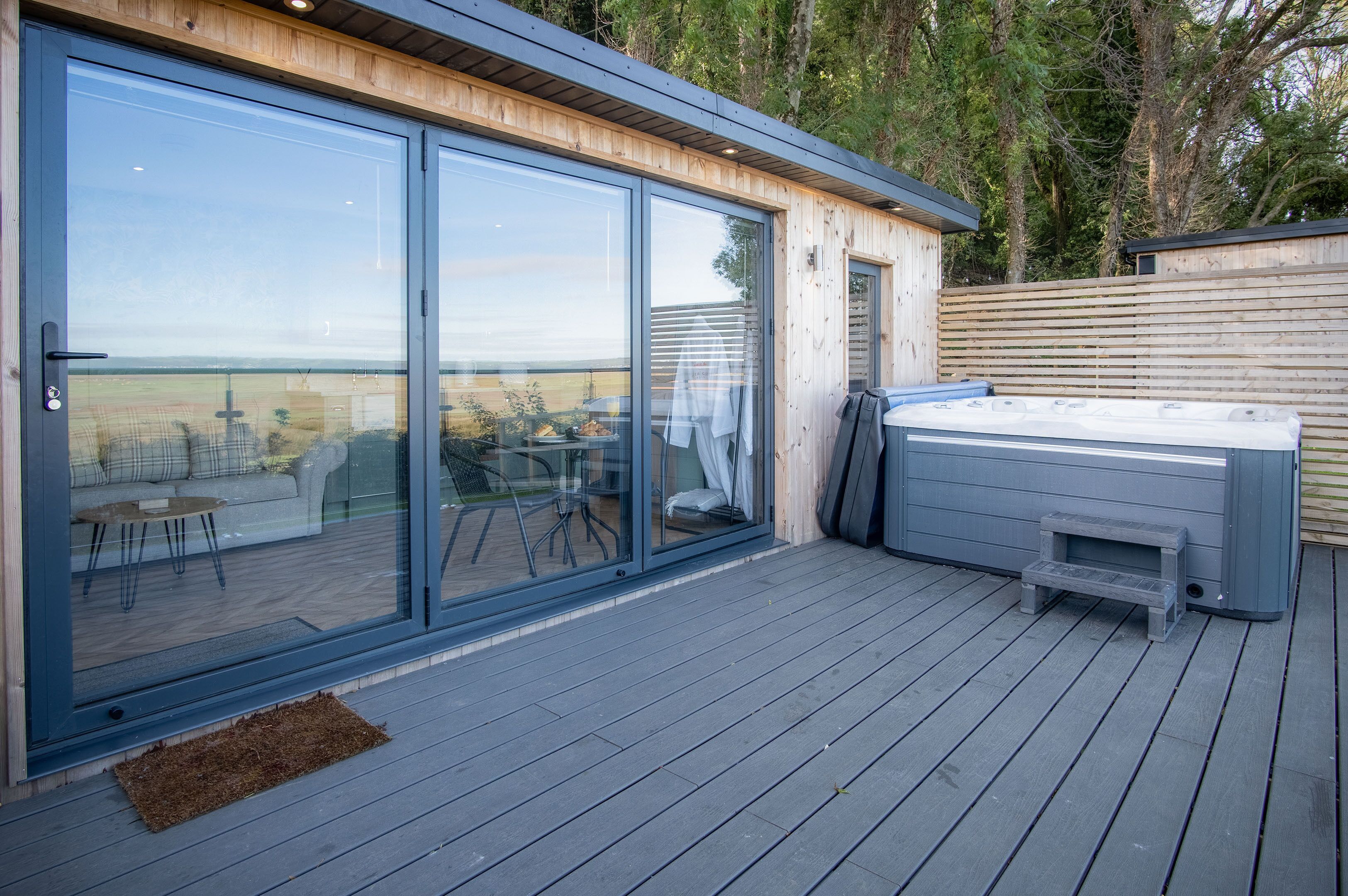 A modern outdoor deck with glass doors leading to a patio area, a hot tub, and a view of the countryside.