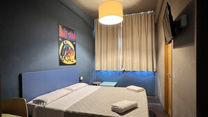 Een hotelkamer met een bed, een bureau, een stoel, een raam met gordijnen, een poster aan de muur en een plafondlamp.
