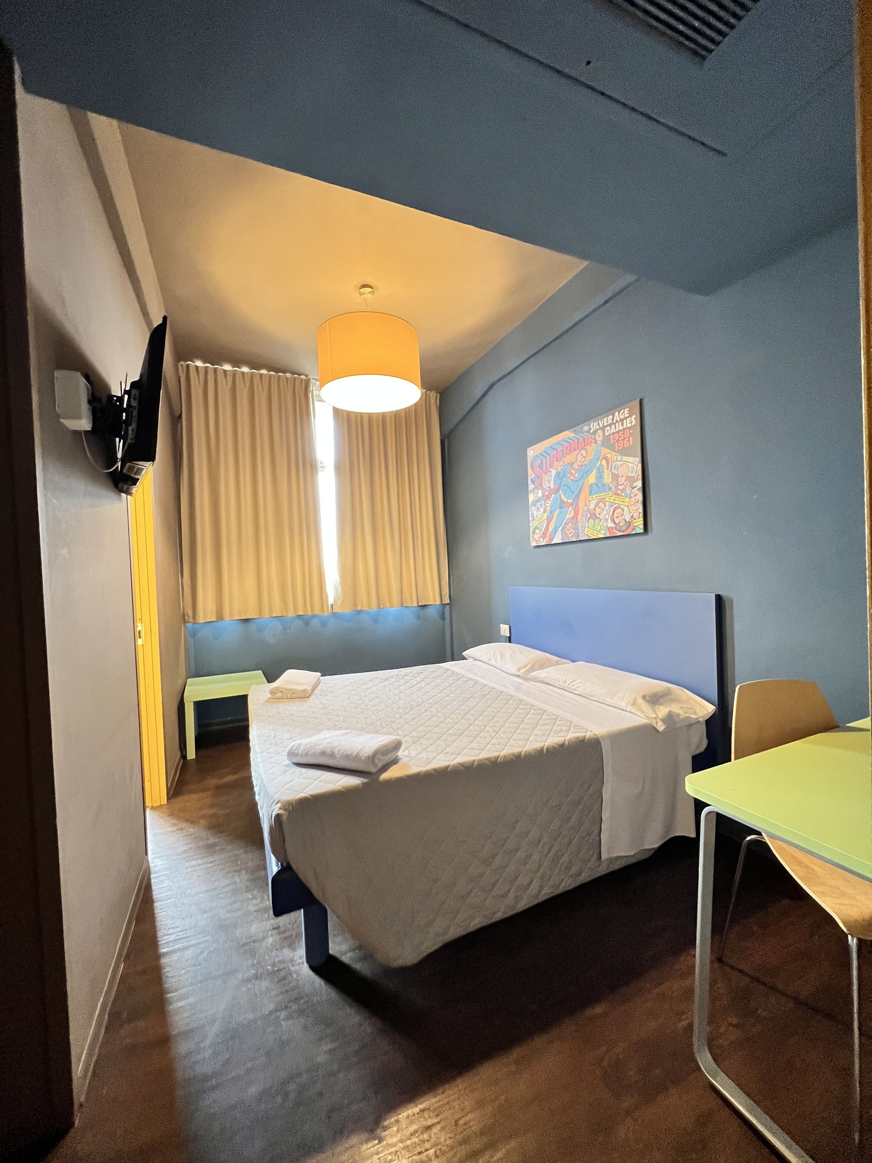 Een hotelkamer met een bed, bureau, stoel, raam met gordijnen, een tv aan de muur en een plafondlamp.