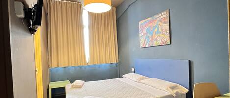A hotel room with a bed, a desk, a chair, a window with curtains, a wall-mounted TV, and a ceiling light.