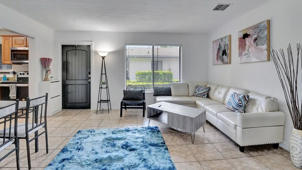 A modern living room with a white sofa, a coffee table, and a blue rug. There is a dining area with a table and chairs, and a kitchen in the background. - Spacious 1 bedroom, East Delray parking, W/D, BBQ (Delray Beach)
