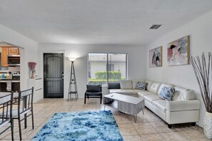 A modern living room with a white sofa, a coffee table, and a blue rug. There is a dining area with a table and chairs, and a kitchen in the background. - Spacious 1 bedroom, East Delray parking, W/D, BBQ (Delray Beach)
