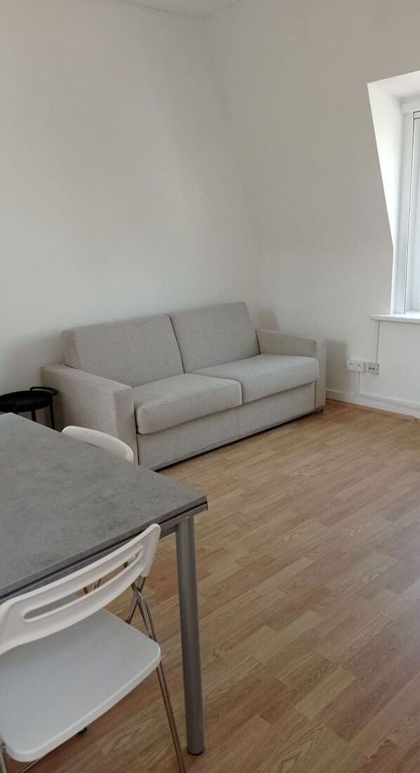 A room with a grey sofa, a table with chairs, and a window with curtains. - Appartement Sympa à 50 m de la mer (Middelkerke)