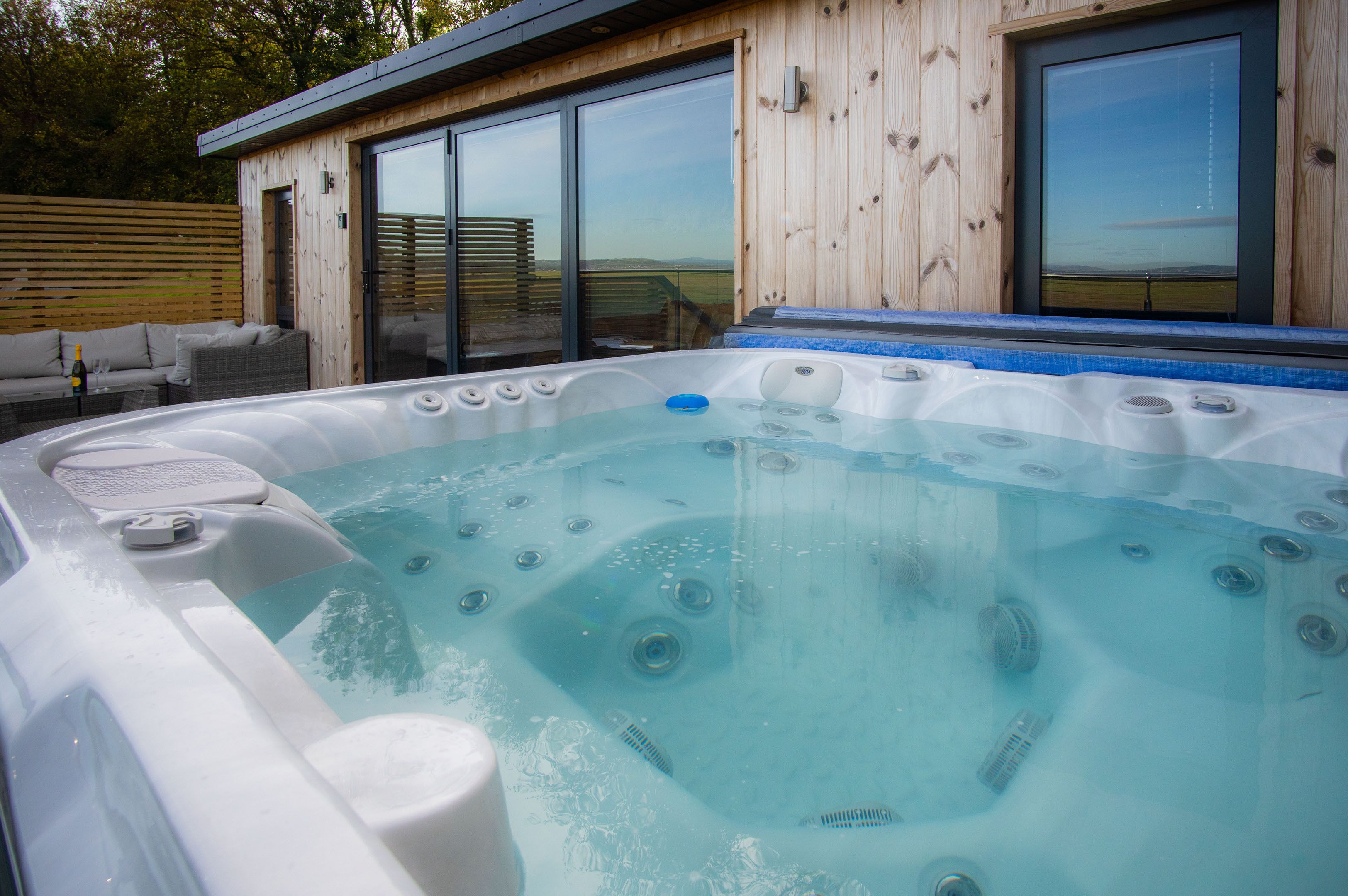 A hot tub with a view of the countryside, a wooden deck, and a glass door leading to a covered outdoor area.