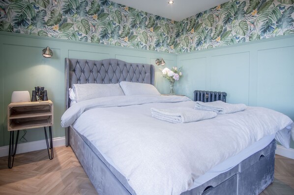 A bedroom with a tufted headboard, a white bedspread, a bedside table with a lamp, and a floral wallpaper. - Rhossili Bay Secret - Cabin - Landimore (Swansea)