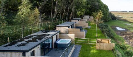 A row of modern cabins with wooden exteriors, a hot tub, and a grassy area with a fence.